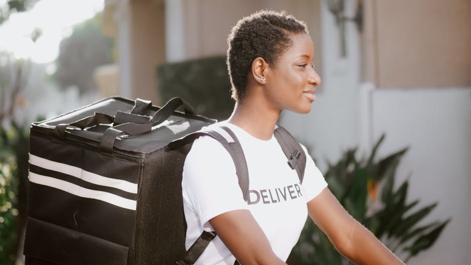 A young woman with short, curly hair and a confident expression, wearing a white t-shirt with 'DELIVERY' printed on it, is seen carrying a large, black insulated delivery bag with white stripes on her