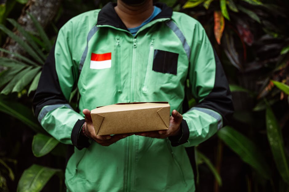 A delivery person in a green and black jacket with a red and white flag patch on the left chest and a black rectangular patch on the right chest is holding a rectangular cardboard box in both hands. T