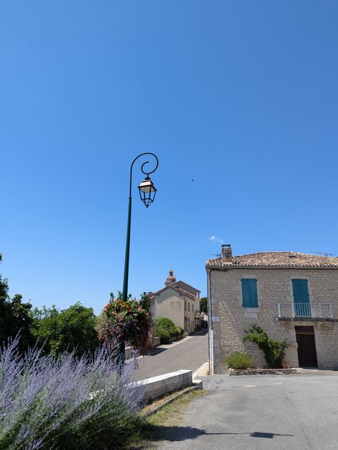 A clear blue sky over a charming street scene featuring a vintage street lamp with an elegant black scroll arm, adorned with a hanging flower basket bursting with pink and purple blooms. To the right,
