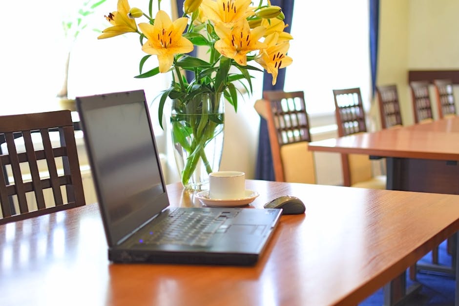 A clear glass vase filled with vibrant yellow lilies and green foliage sits on a polished wooden conference table in an office setting. The bouquet features large, open blooms with prominent stamens, 