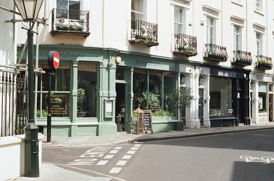 A storefront of a florist shop on a city street corner, featuring large glass display windows filled with vibrant floral arrangements. The shop's exterior is painted in a pale green hue with decorativ
