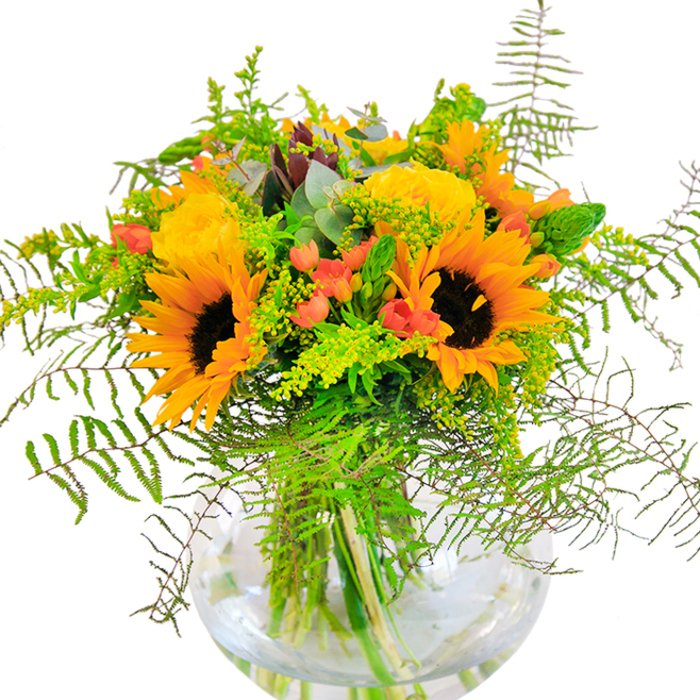 First impressions are radiant as this bouquet greets you-a sun-drenched mix of yellow roses and towering sunflowers arranged in a round, crystal-clear vase, like a piece of summer brought in from Barnes Pond. Each sunflower beams outward, petals glowing cheerfully around a mahogany centre, and is joined by garden roses whose layered petals swirl in buttery softness and subtle fragrance. Dotted through the arrangement, rich red Hypericum berries add a sparkle-tiny orbs of colour that play against the luxurious yellows and deep, earthy greens. Feathery fronds of fern and graceful vine-like greenery spill gently over the vase's rim, providing a breezy sense of movement akin to the windswept hedgerows that line Barnes' quiet lanes. Golden sprigs-perhaps solidago-offer subtle clusters of miniature yellow blossoms, complementing the sunny palette, while eucalyptus leaves punctuate the bouquet with cool blue-green hues and a faintly invigorating scent. The round vase, reminiscent of classic English glassware, holds the display together, allowing stems to be glimpsed in clear water and enhancing the freshness of the whole. This arrangement conjures memories of summer fetes and garden parties by the river-a fitting gift for joyful occasions or welcoming friends into a Barnes home. Every detail speaks of local artistry and warmth, reflecting the expertise of your trusted neighborhood florist.